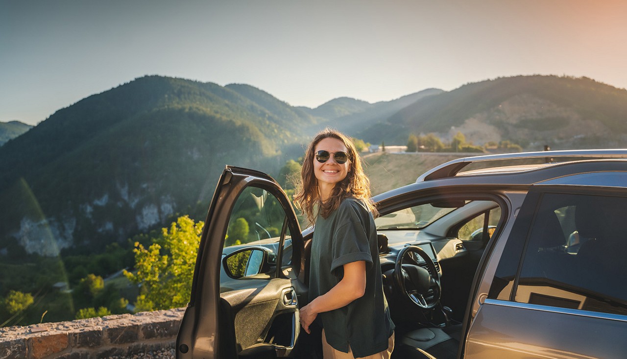 A smiling young woman stepping out of her car near a scenic lookout point overlooking a beautiful mountain and valley.