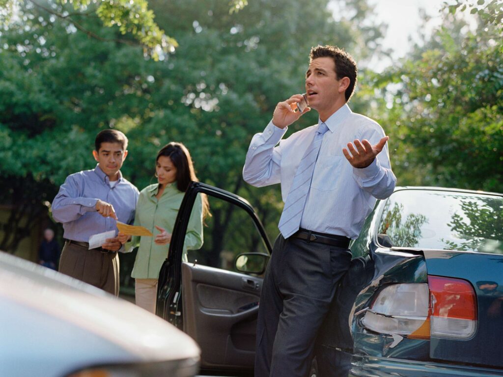 A man talking on a phone while a couple look up their insurance after they both collided in an accident.